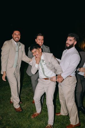 Groomsmen in light colored suits share a playful moment during outdoor wedding celebrations.
