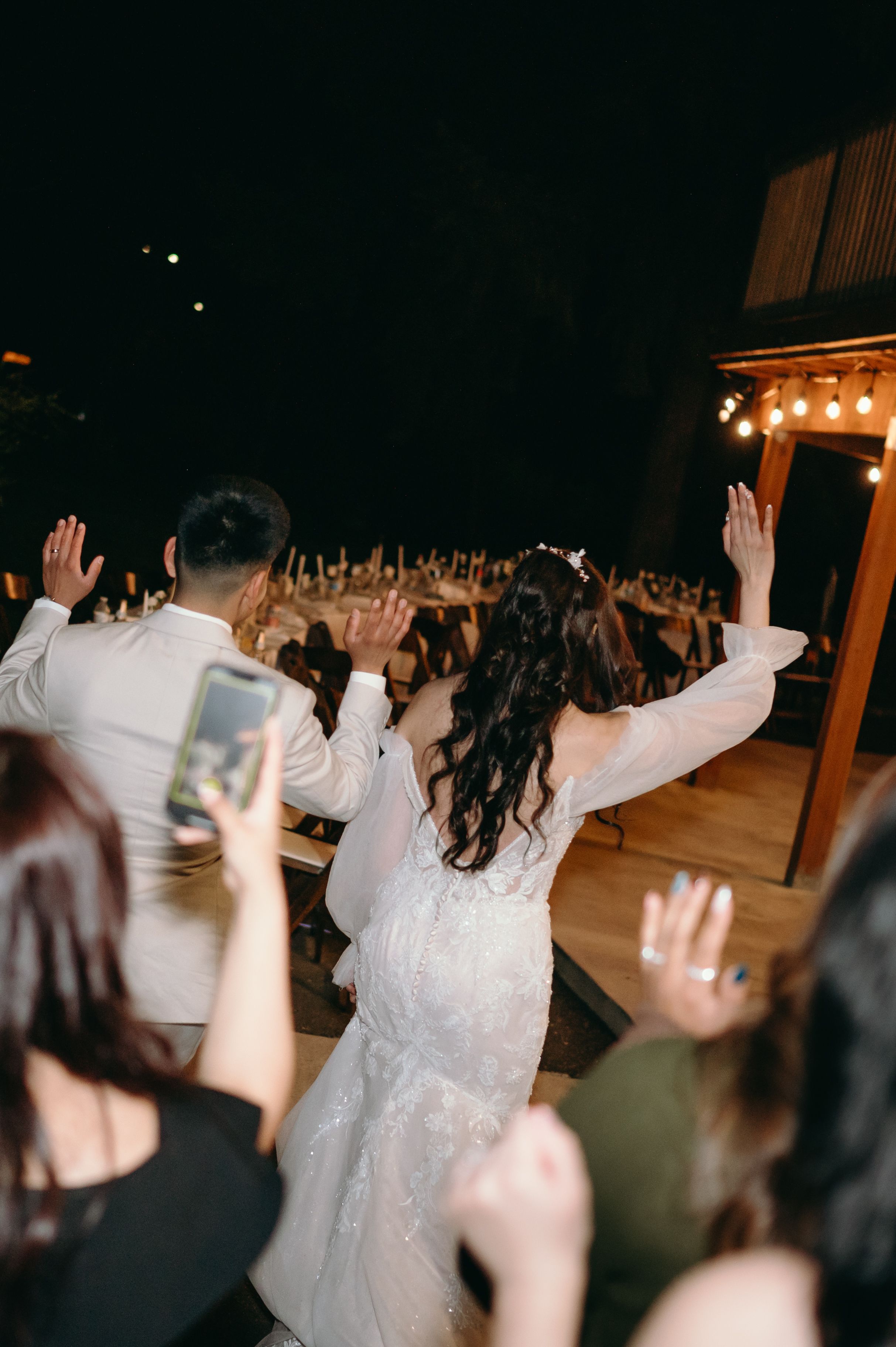 Newlyweds raise their arms in celebration while facing their reception crowd.