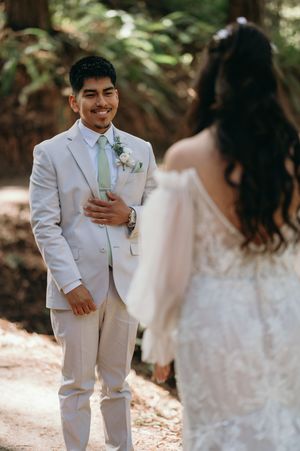A groom in a light beige suit with mint tie smiles joyfully during a first look moment with his bride in the woods.