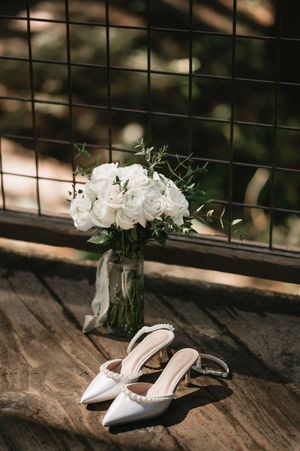 Elegant white bridal bouquet in glass vase with ivory slingback heels on rustic wooden surface.