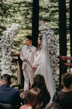 A couple stands under a white floral arch during their forest wedding ceremony.