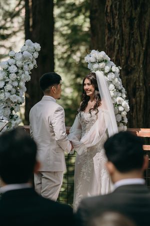 A couple shares their wedding ceremony under white floral arches in a romantic forest setting.