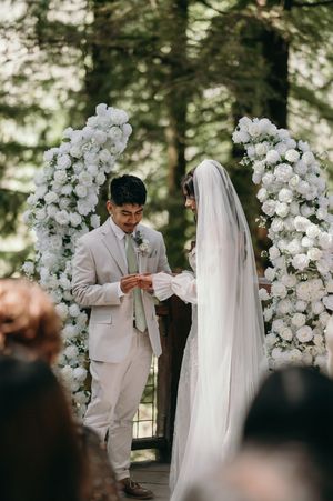 A wedding ceremony under a white floral arch in a forest with guests watching.