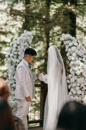 A wedding ceremony under an arch of white roses in a woodland setting with soft natural lighting.