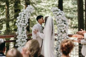 An outdoor wedding ceremony under beautiful white floral arches with guests seated in a forest setting.