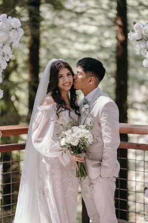 A romantic moment captured between newlyweds under white floral arches in a forest setting.