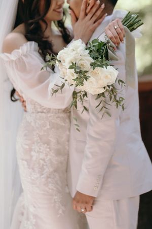 Close-up of wedding couple holding white rose bouquet with greenery.