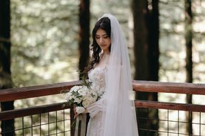 White-veiled bride in a lace dress holds bouquet while standing on wooden deck in forest setting.