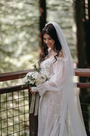 Bride in lace gown with flowing sleeves poses on forest deck with white bouquet.