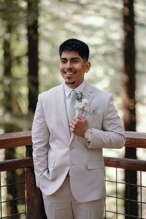Wedding attire featuring a light beige suit and boutonniere in a forest setting.