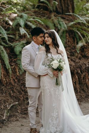 Wedding couple embraces on a forest trail while the bride holds a white floral bouquet against a backdrop of ferns.