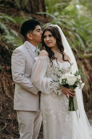 Romantic wedding portrait of a couple in light colored attire surrounded by forest greenery.