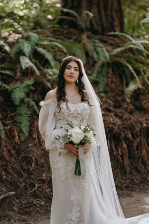 Bride in an elegant lace dress holds a white bouquet while standing on a forest trail.