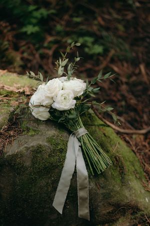 An elegant white rose and greenery wedding bouquet resting on a mossy rock with satin ribbon streamers.