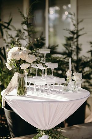 White floral centerpiece and champagne tower setup on a draped table with evergreen backdrop.