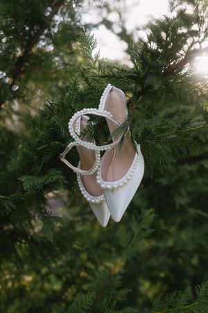 Elegant white bridal shoes with pearl detailing hanging on evergreen branches in soft natural lighting.