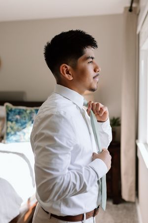 A person in a crisp white button-down shirt adjusts their tie while looking thoughtfully out a window.