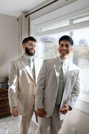 Two people in light beige suits and ties smiling warmly by a window.