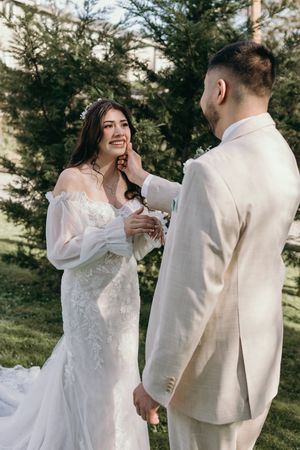 A touching wedding moment between newlyweds in white attire against an evergreen forest backdrop.