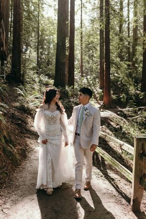 A romantic wedding couple walks hand in hand along a sunlit forest trail surrounded by towering redwood trees.