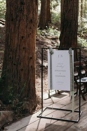 Wedding welcome sign displayed on metal stand among redwood trees.