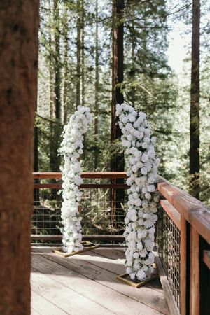 White floral arch pillars decorate a wooden deck surrounded by tall redwood trees.