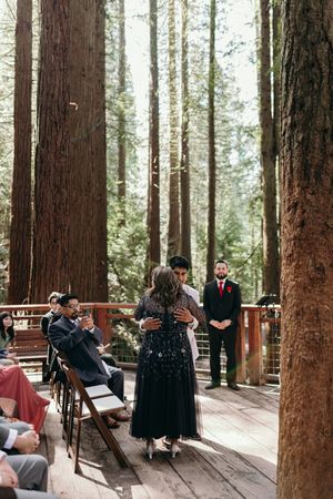 An intimate forest wedding ceremony takes place on a wooden deck surrounded by towering redwood trees.