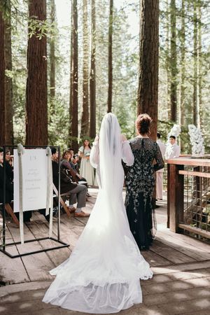 A wedding ceremony setup on a wooden deck in a forest with tall redwood trees and white decor.