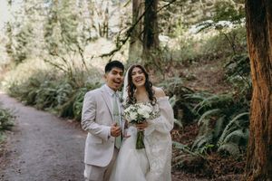 A wedding couple poses on a forest path surrounded by lush ferns and tall trees.