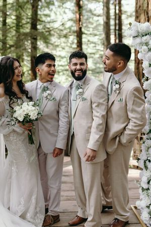 Wedding party in beige suits and white gown share joyful laughs during outdoor forest ceremony with floral decorations.