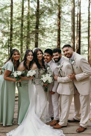 Wedding party in beige suits and sage green dresses pose together in a forest setting.