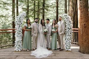 A wedding party stands between white floral arches on a wooden deck in a forest setting.