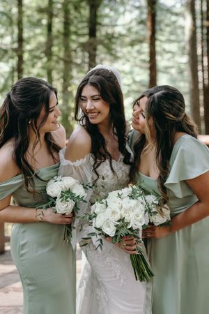 Bridesmaids in sage green dresses share a candid moment with the bride in a forest setting.