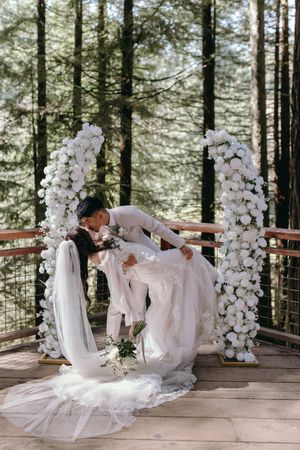 Dramatic wedding dip pose under floral arches on a wooden deck in a forest setting.