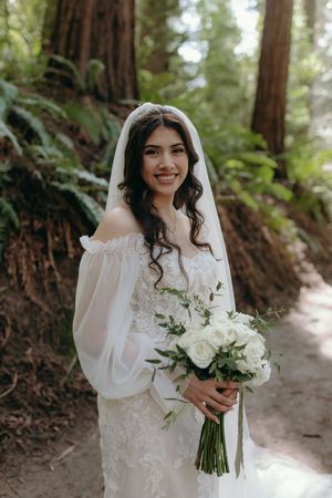 Bride holding white bouquet poses in off-shoulder gown against redwood forest backdrop.