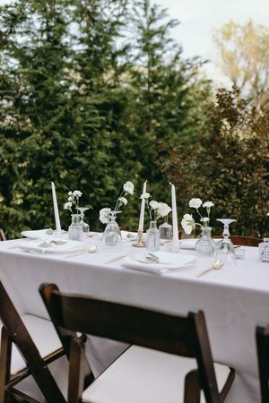 A romantic outdoor table setting with white linens, taper candles and small floral arrangements in glass bud vases.