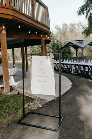 Wedding welcome sign displayed on a metal stand beneath a wooden deck with string lights.