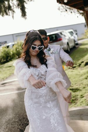A bride wearing a white lace gown with puff sleeves and sunglasses poses outdoors on a sunny day.