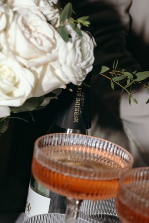 Elegant cocktail glass with orange drink next to white roses and greenery.