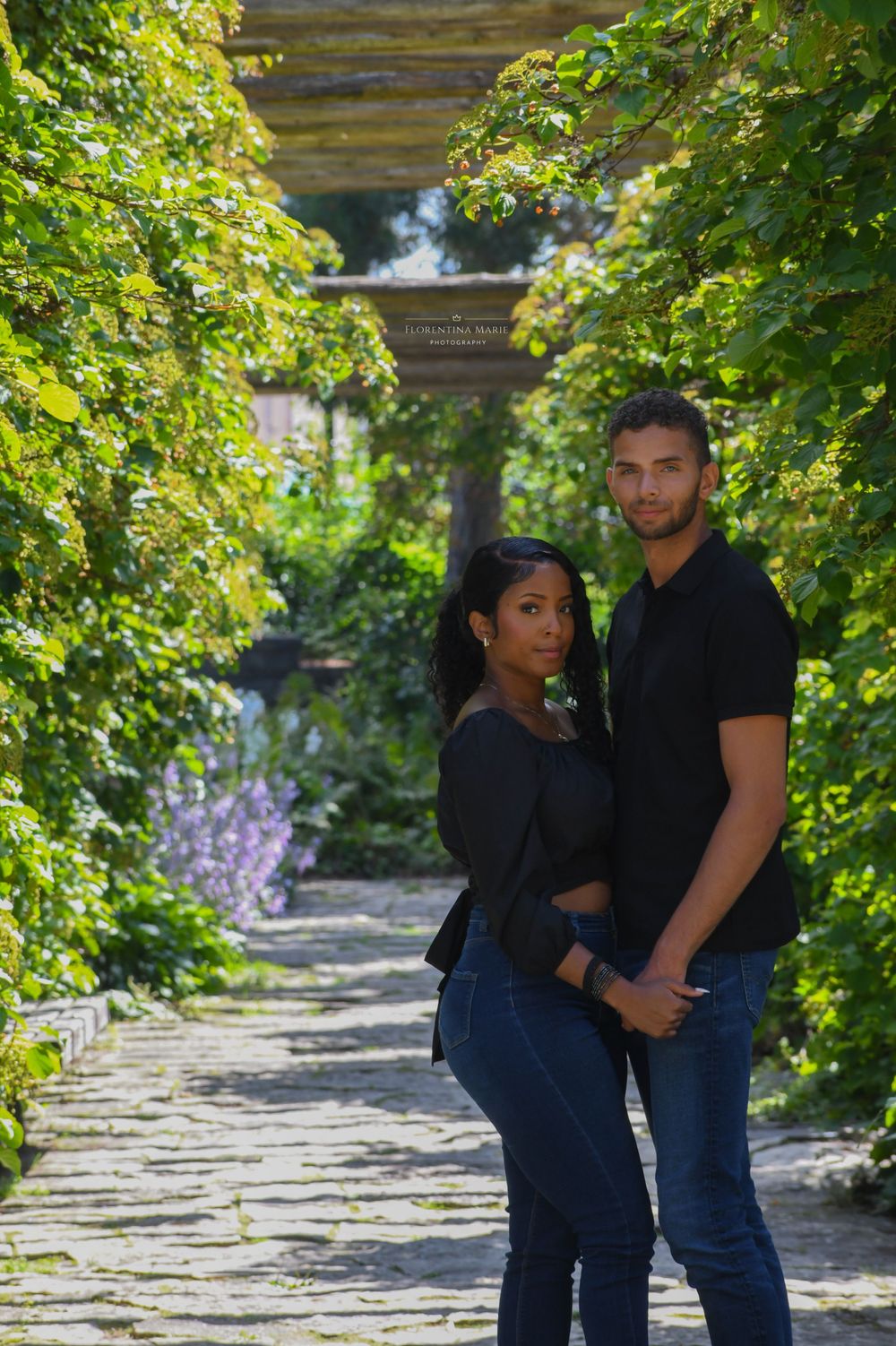 Couple poses together on a stone path under a pergola with blooming purple flowers in the background.