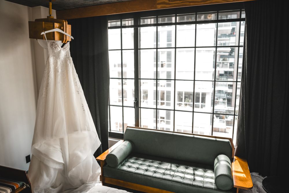 Wedding dress hanging in window with natural light streaming through dark room interior.