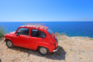 Vintage red Fiat 600 parked on coastal cliff overlooking blue Mediterranean Sea.