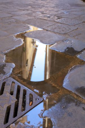 Puddle reflection of building on wet cobblestone street with drain grate.
