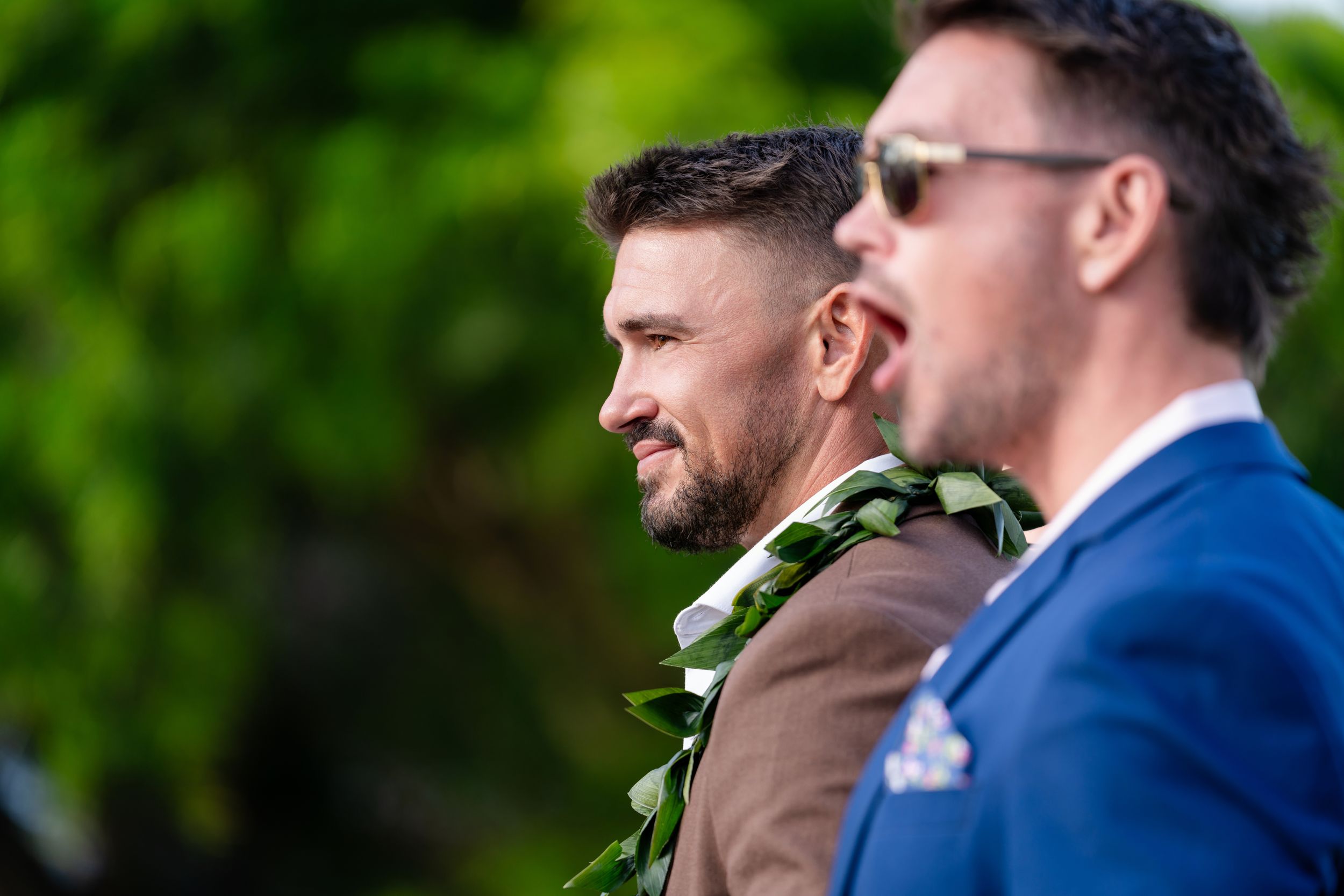 Two men in formal attire wearing floral leis at an outdoor Hawaiian wedding ceremony.