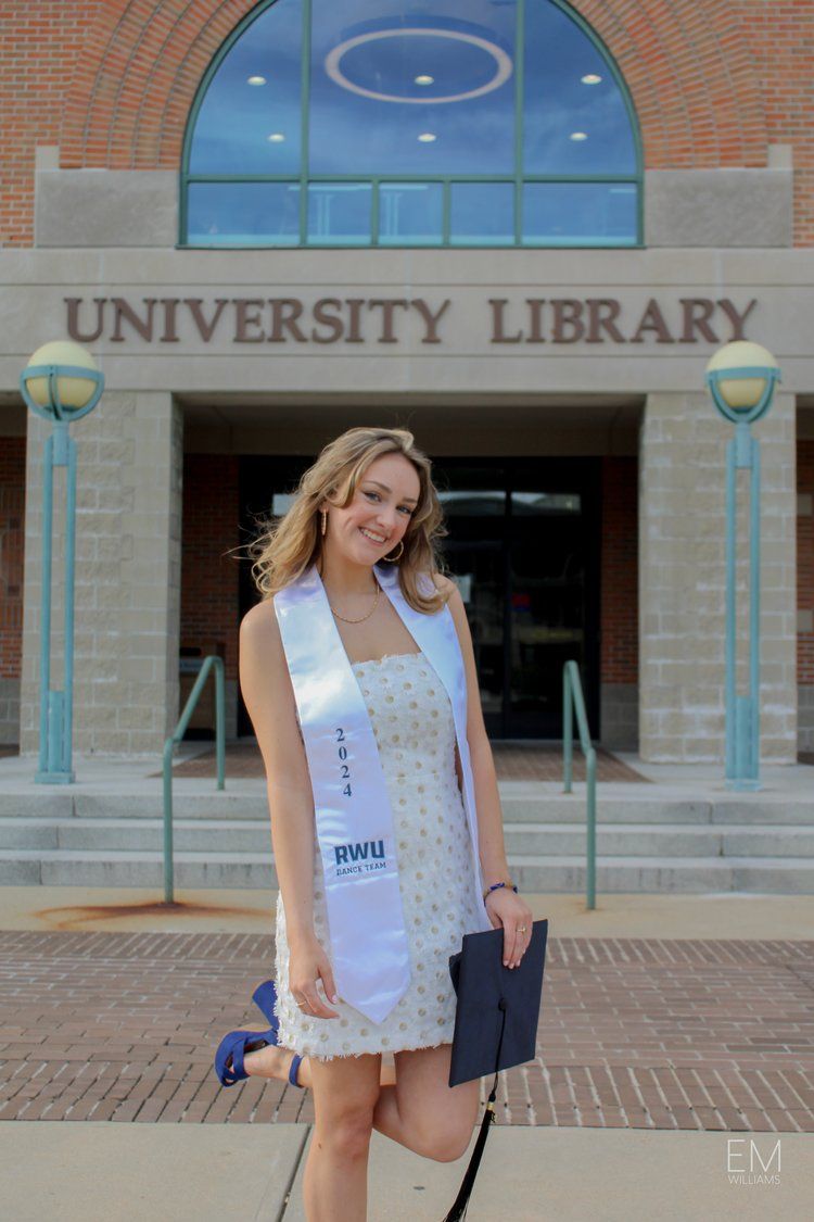 A graduate poses in academic regalia outside a brick university library entrance.