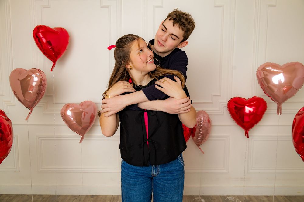 Playful couple embracing against white wall with red and pink heart balloons.