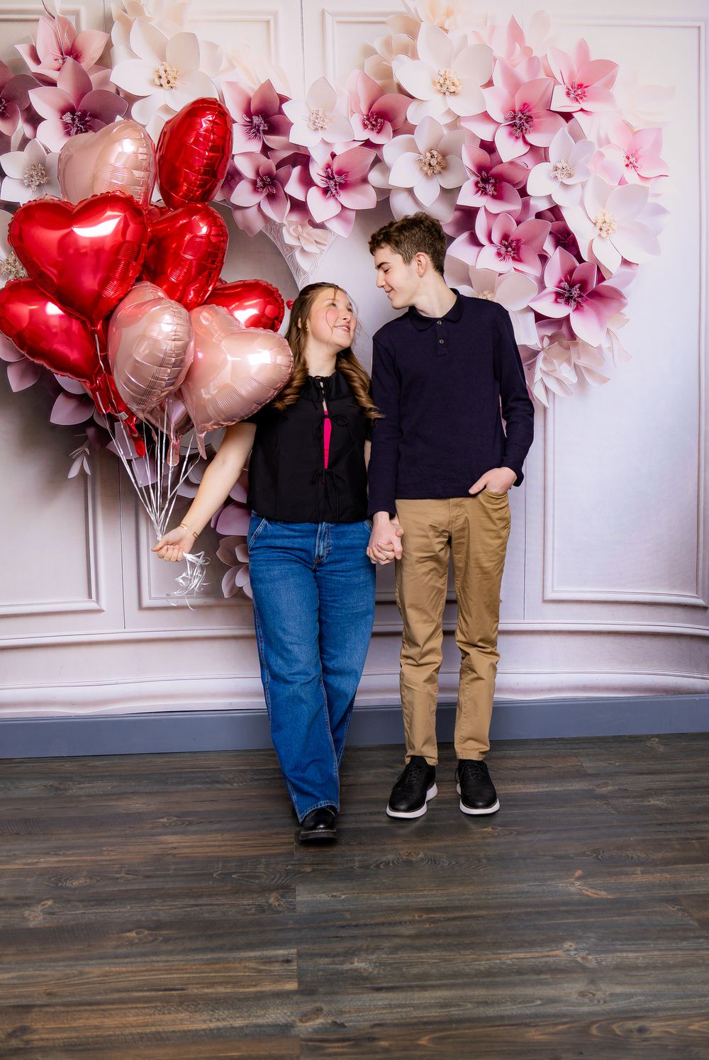 Romantic couple pose with heart balloons and pink flower wall backdrop.