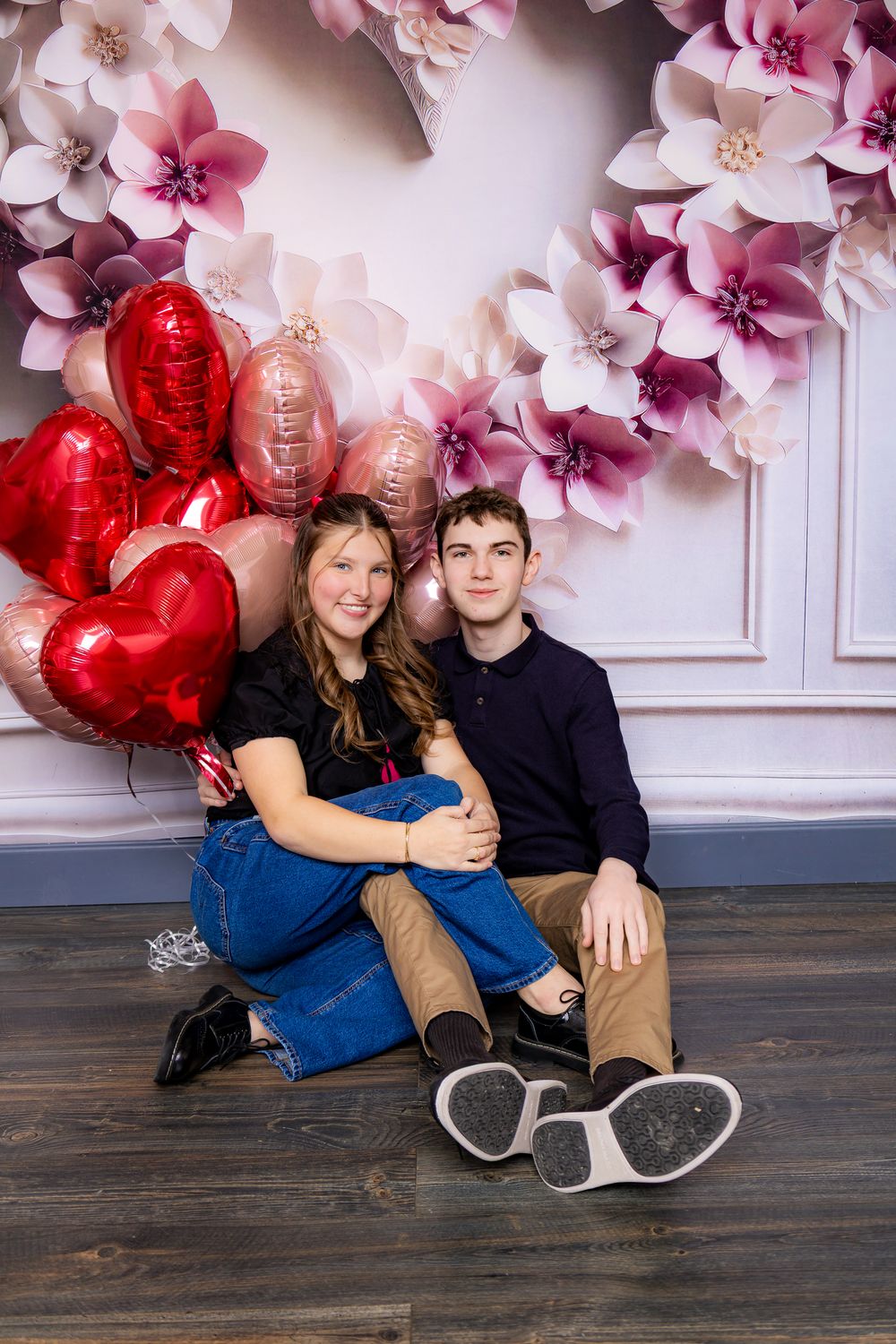 Romantic Valentine's photo with pink floral arch and red heart balloons against white wall on wooden floor.