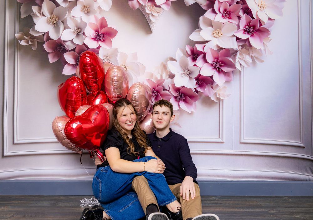 Couple sitting together against pink floral backdrop with red and rose gold heart balloons.