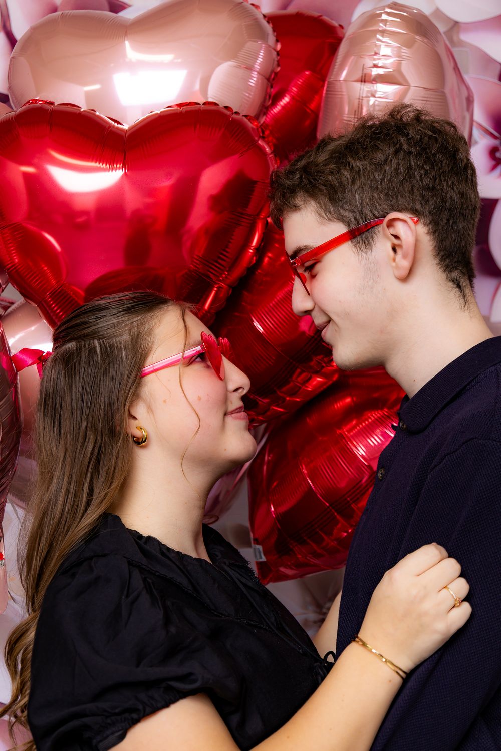 Romantic couple moment against pink floral wall with red heart balloons in background.